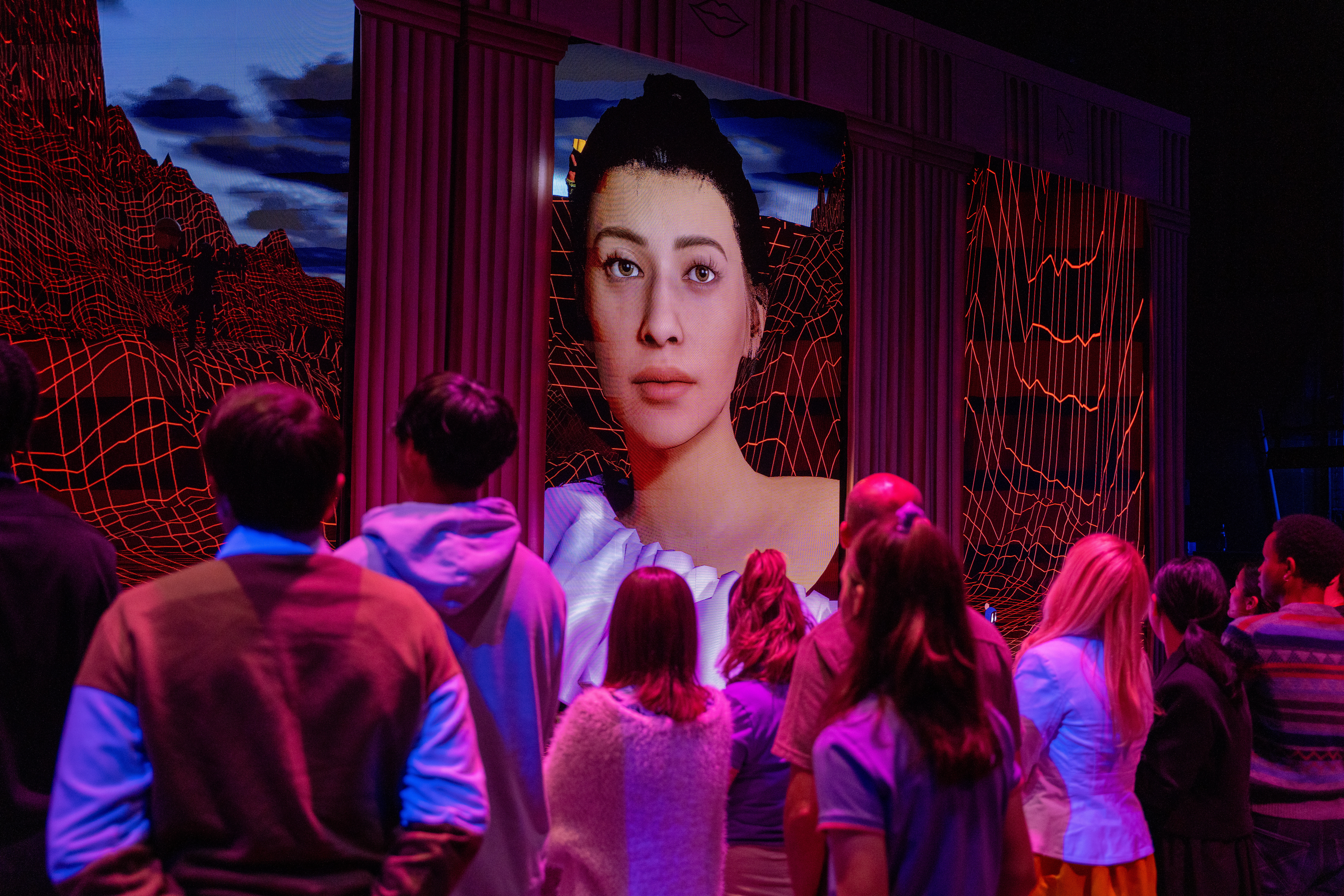 Theater goers on a stage look at a large screen with an AI avatar