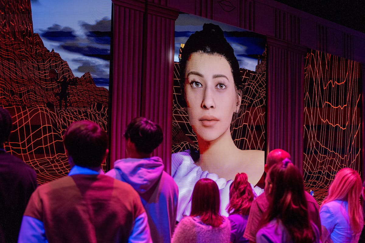 Theater goers on a stage look at a large screen with an AI avatar
