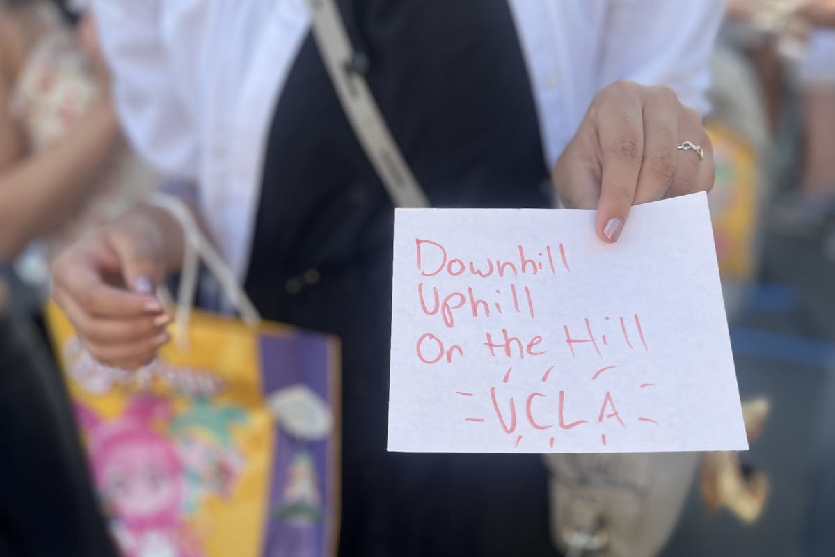 A person holds a handwritten sign toward the camera that reads "downhill, uphill, on the hill, ucla"
