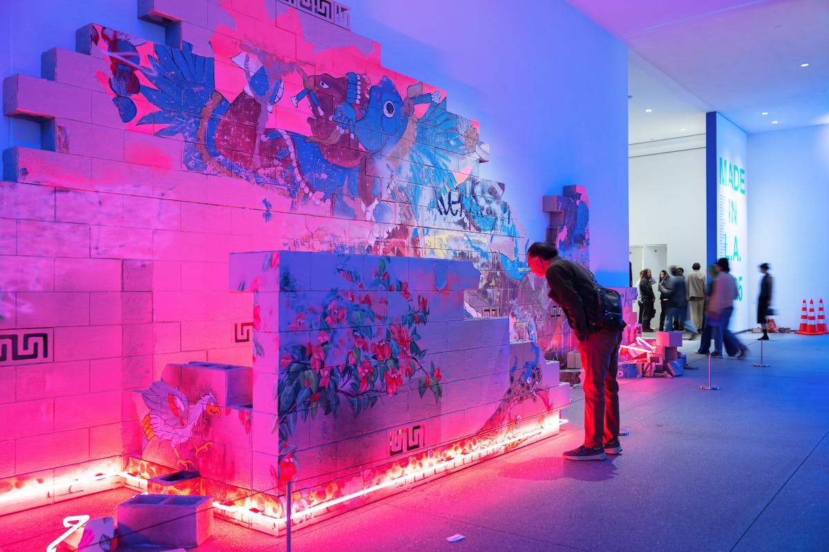 A person looks at a large scale work installed at the hammer museum, made from bricks, neon paint and pink neon lights.