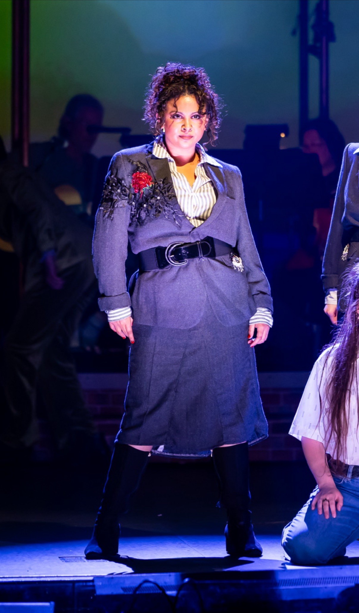 A female performer on stage looks dramatically into the audience. She is wearing a grey suit with a red flower on the lapel