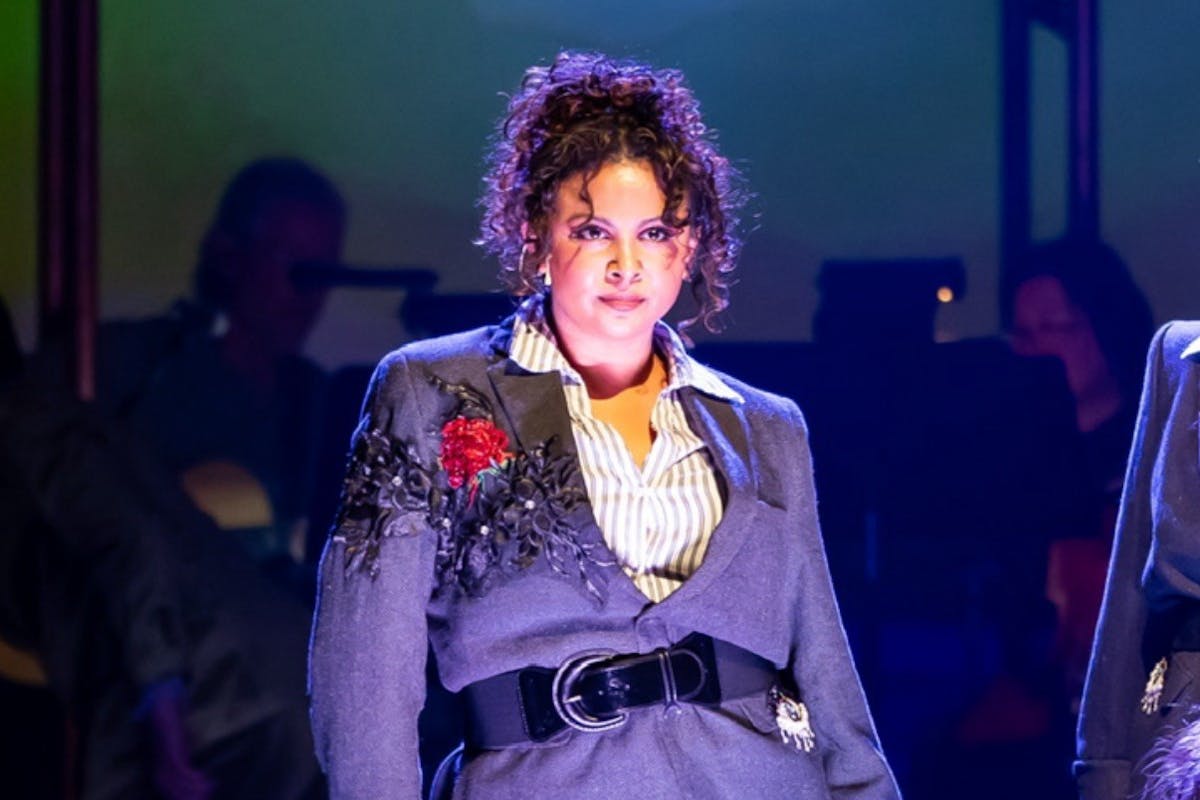 A female performer on stage looks dramatically into the audience. She is wearing a grey suit with a red flower on the lapel