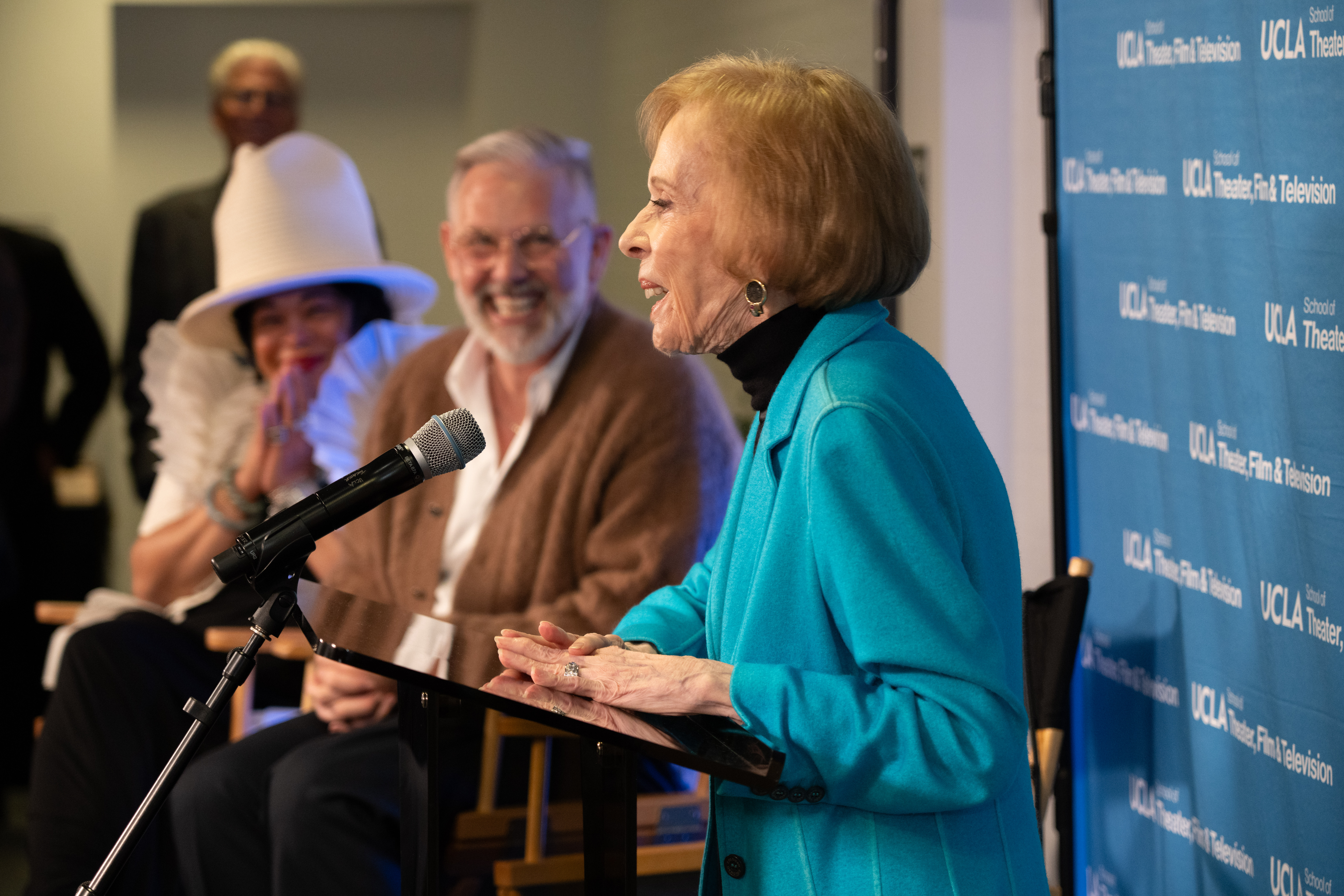 carol burnett speaks at a podium while onlookers smile