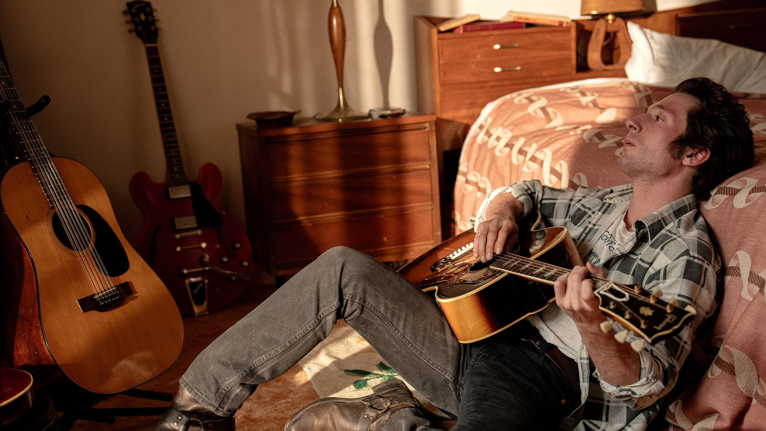  a man holding a guitar leans back against the side of a bed, deep in thought