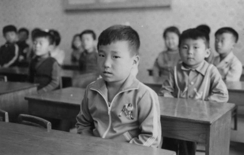 young boy sitting at a desk in a classroom with other children in the background