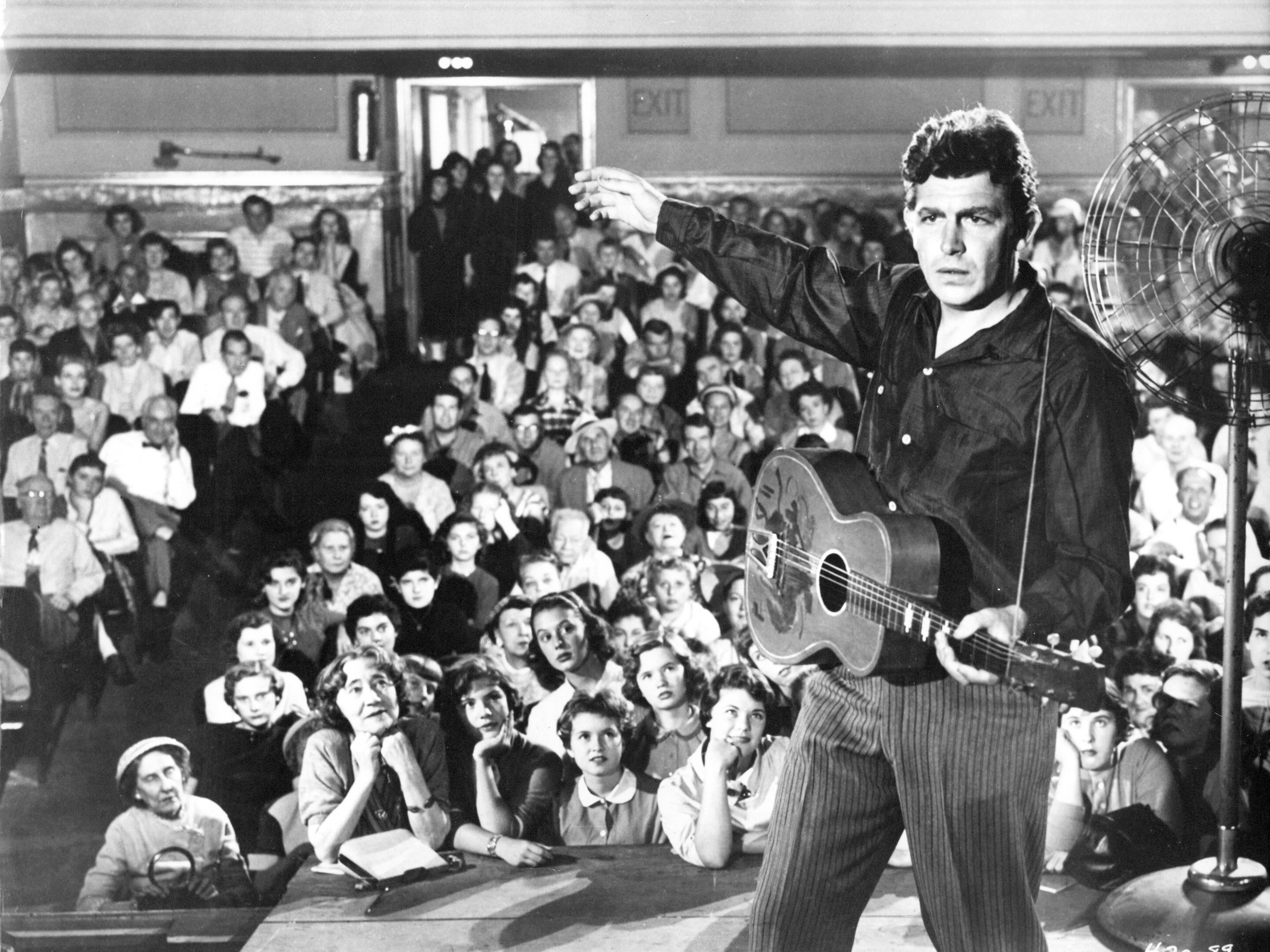 Man with guitar on a stage with audience behind him