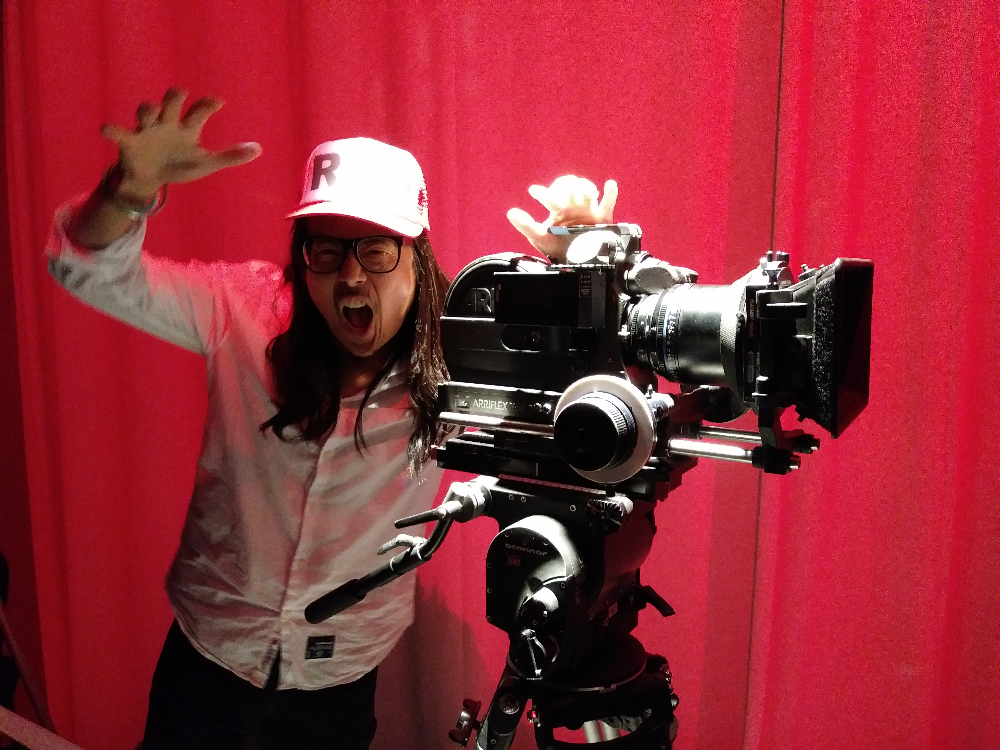 Jon Moritsugu making faces with arms upraised while standing behind a large camera 