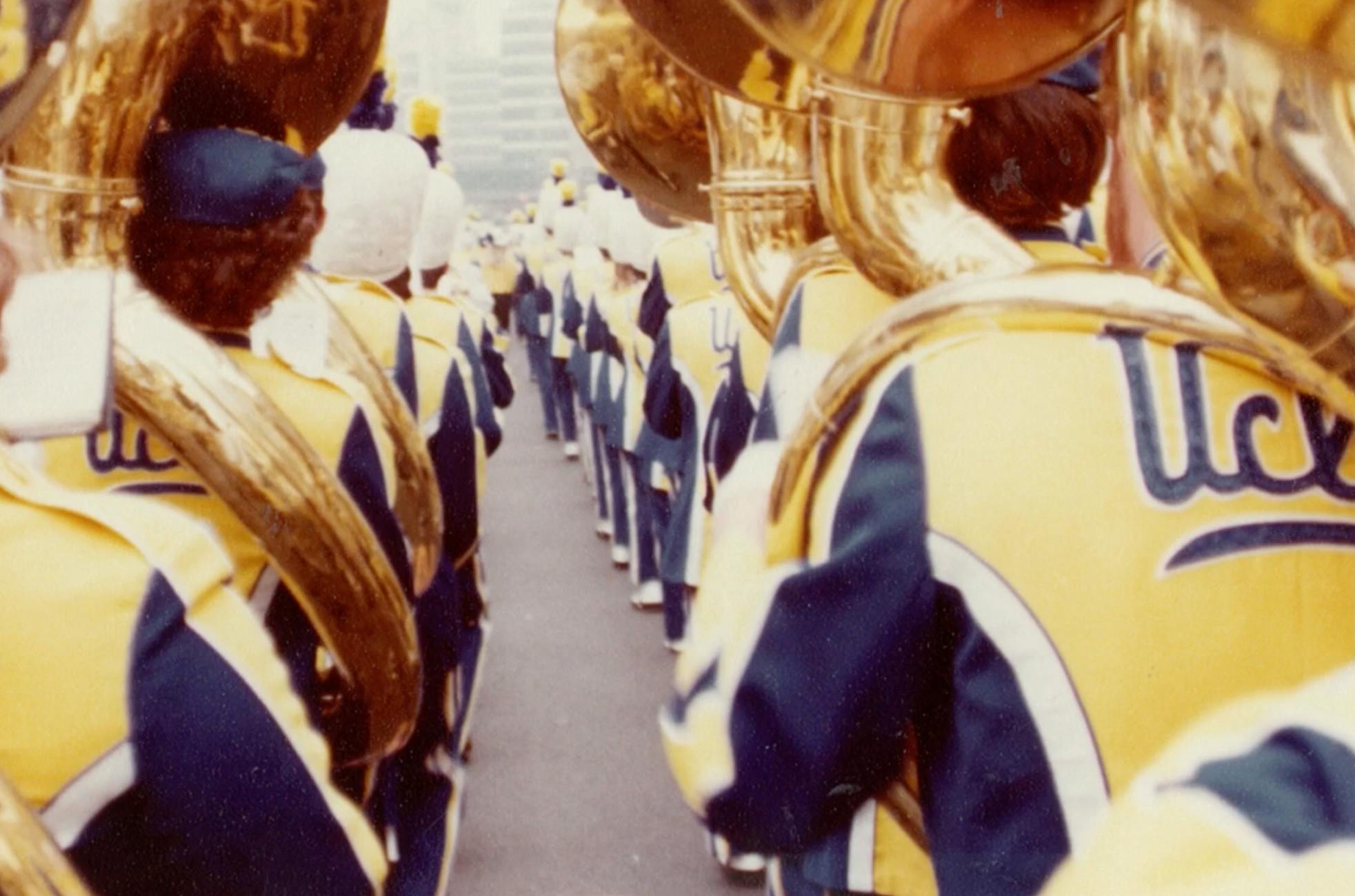 Shot of UCLA marching band in a row from behind. The back of tubas and vintage uniforms can be seen.