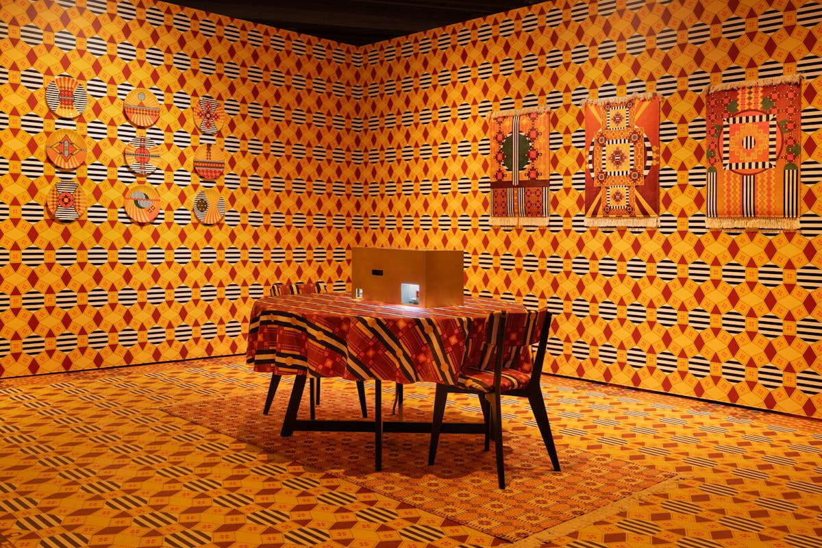 An art installation made striking by a wash of orange patterns on a floor, two walls a table and art hanging on the orange walls