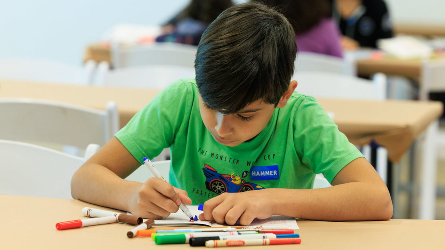 A young boy in a green shirt writes with colorful markers