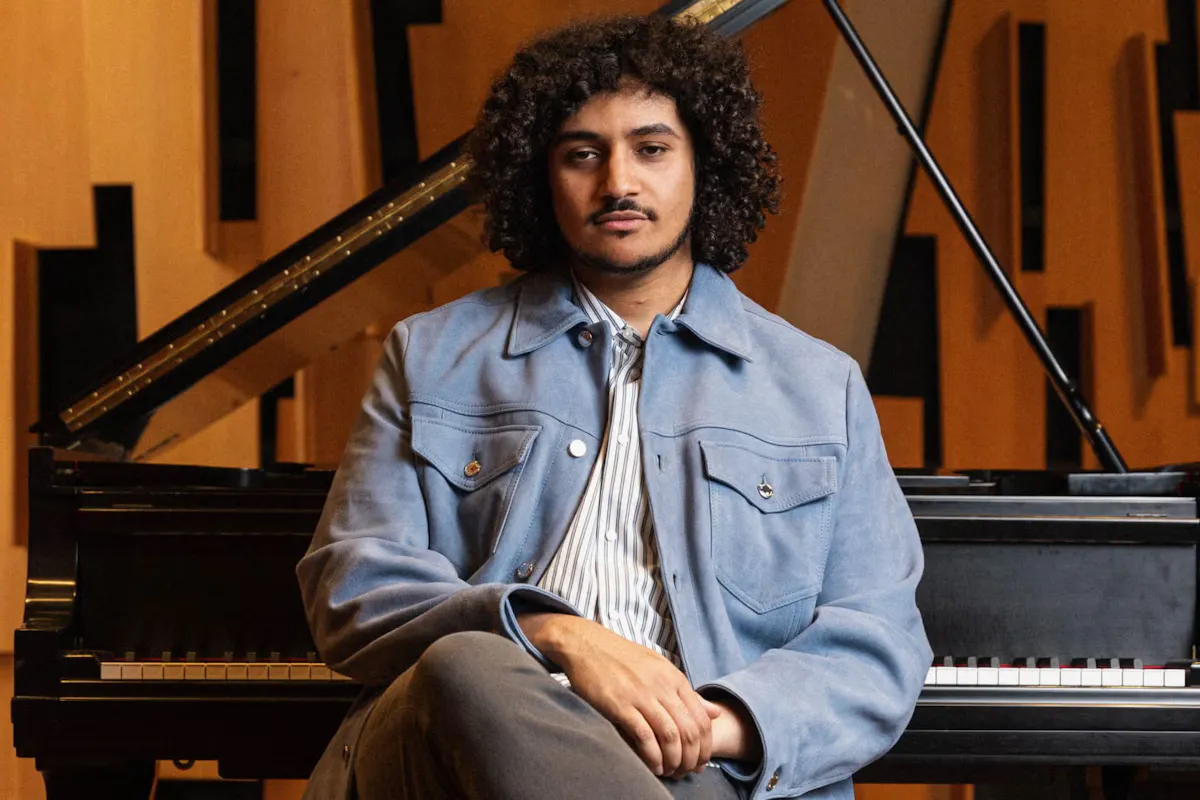 Composer Naveed Perkins sits in front of a piano with his legs crossed
