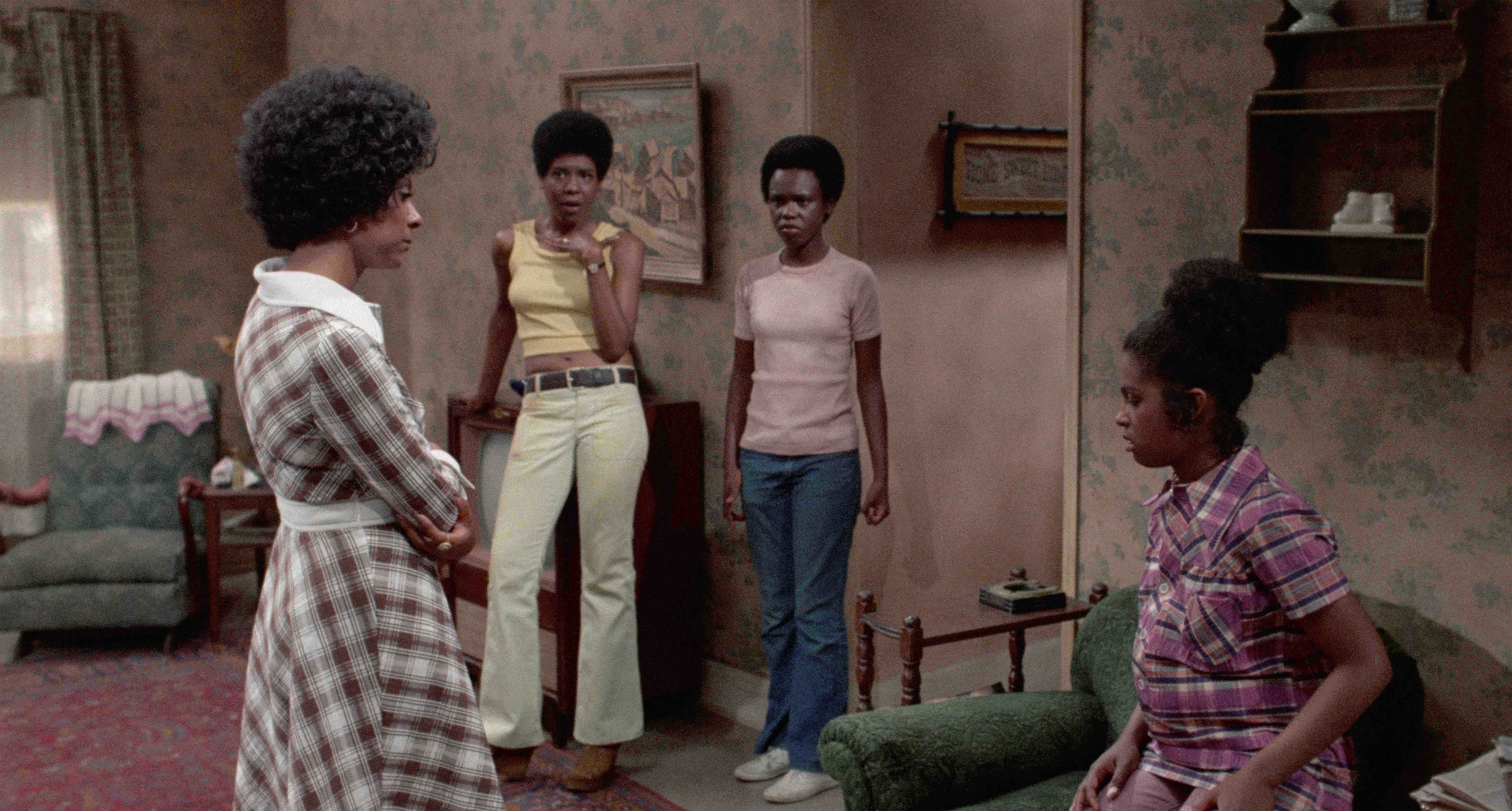 three women standing in a living room while one sits