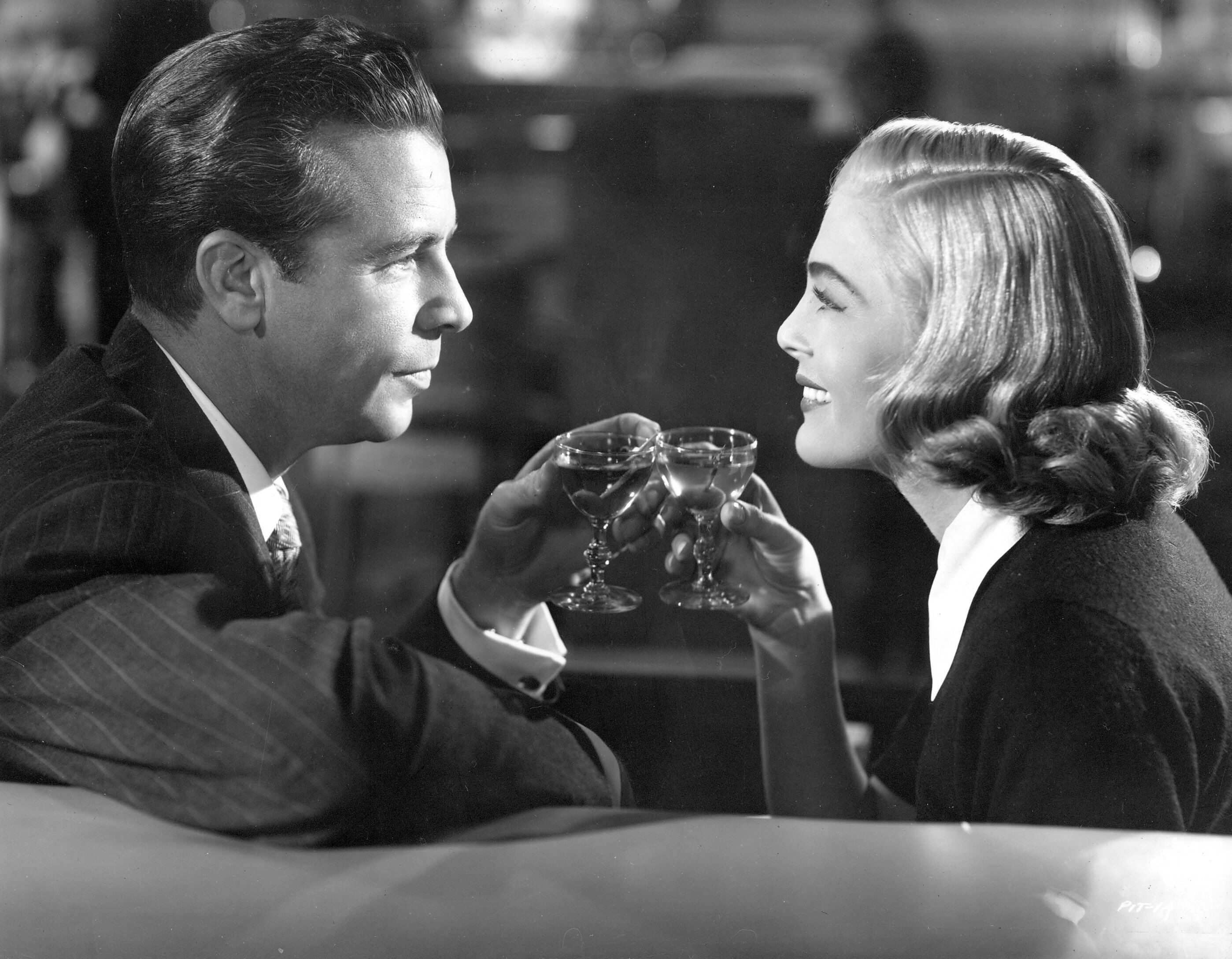 Man and woman seated holding small glasses of drinks and looking at each other while man stares and woman smiles