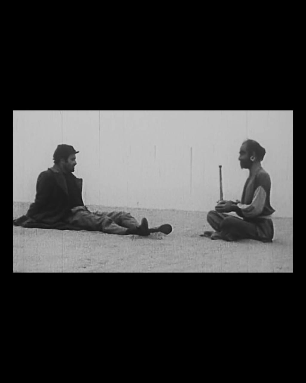 Man reclining on ground facing another man sitting cross-legged