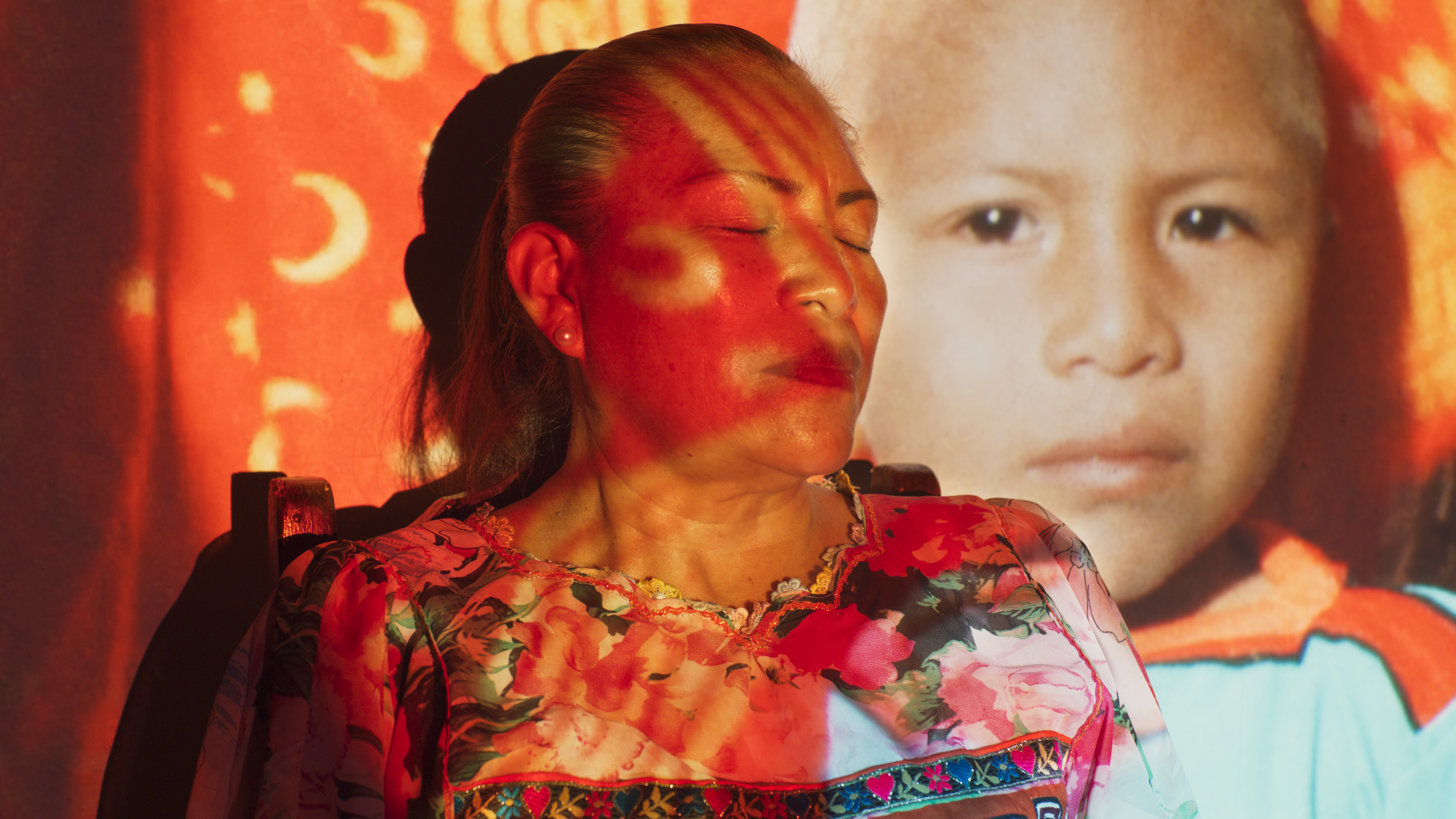 Dramatic screen shot from a film. A woman stands in front of a screen with her eyes closed while an image is projected onto her and the screen. 
