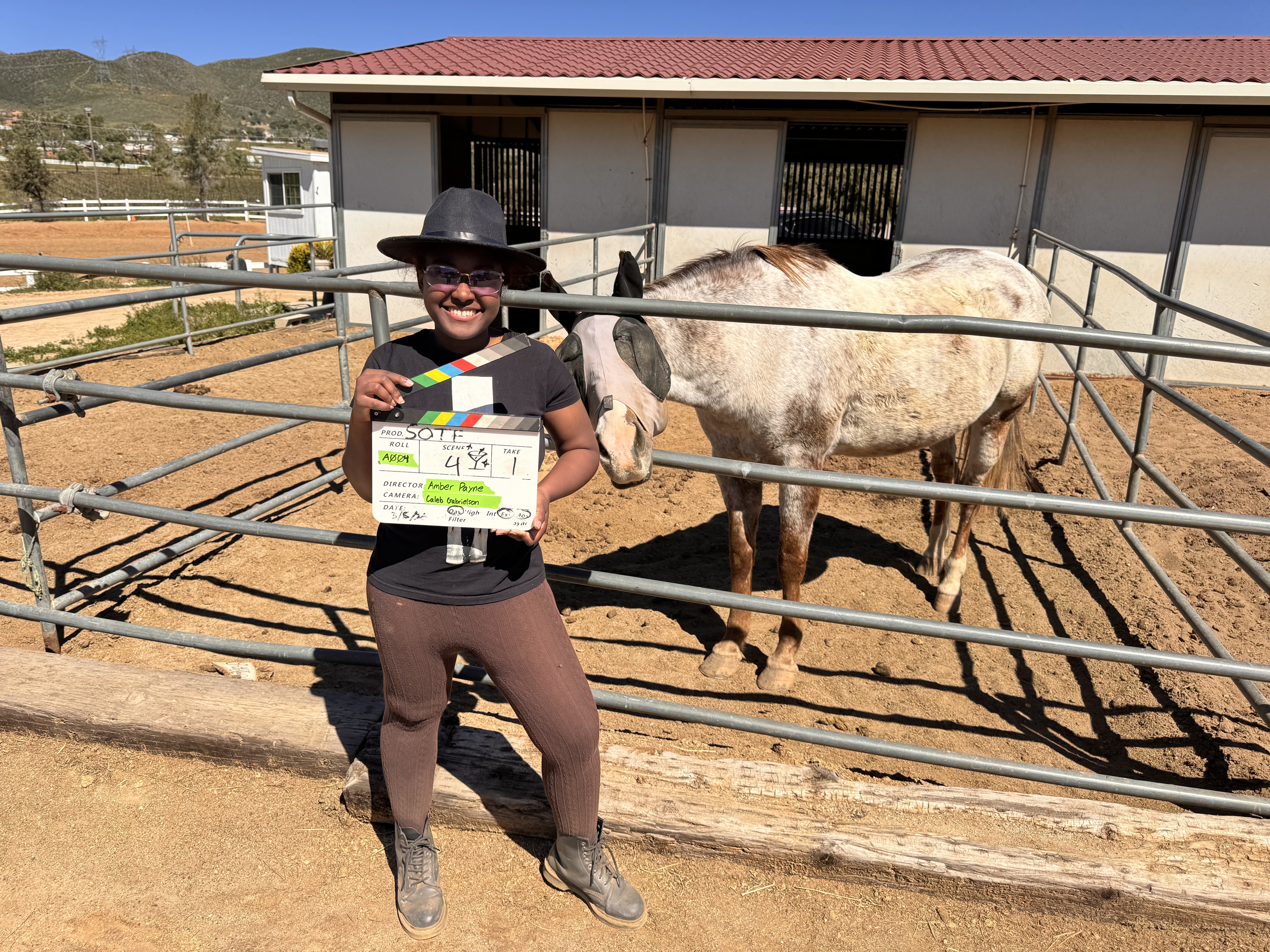 A young filmmaker on a set with a horse corral and white horse holds a scene clapboard
