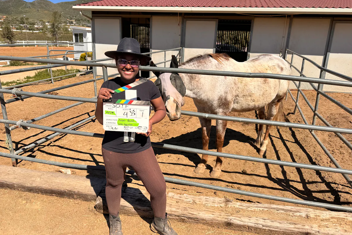 A young filmmaker on a set with a horse corral and white horse holds a scene clapboard