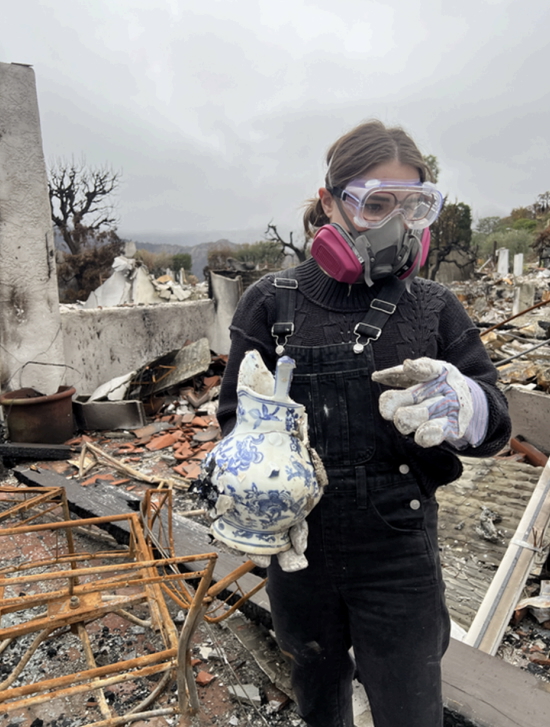 A young woman wearing protective gear sorts through the rubble of a fire.