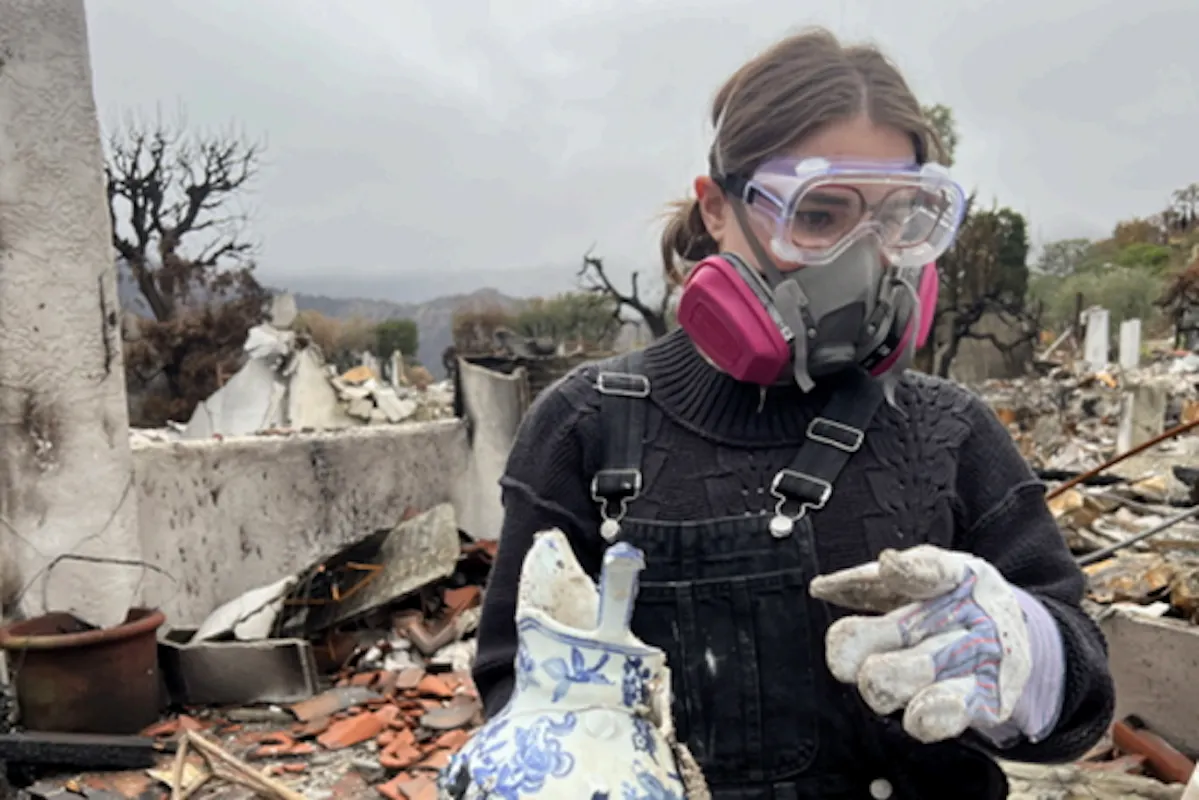 A young woman wearing protective gear sorts through the rubble of a fire.