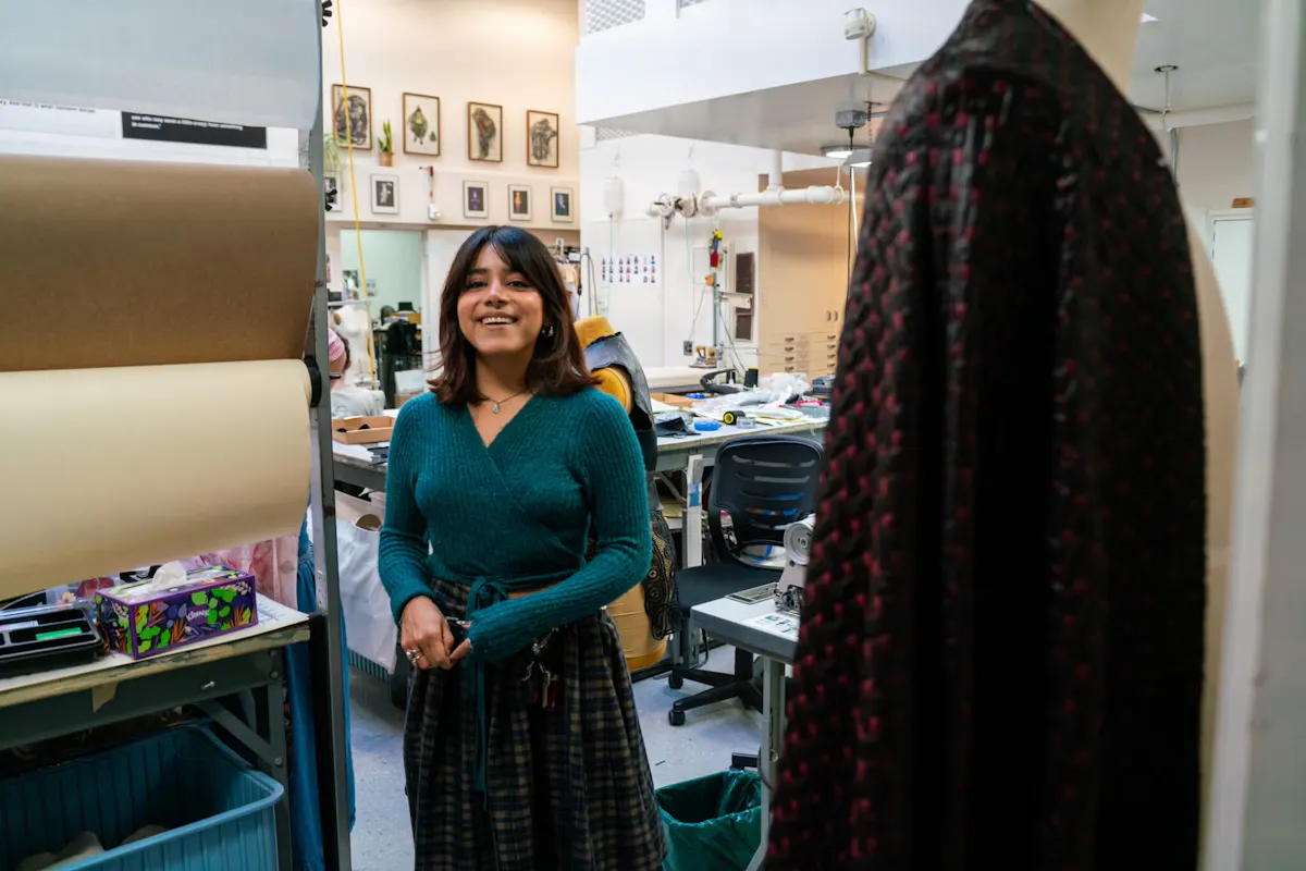 A young woman stands at the door of a room filled with sewing machines and fabric