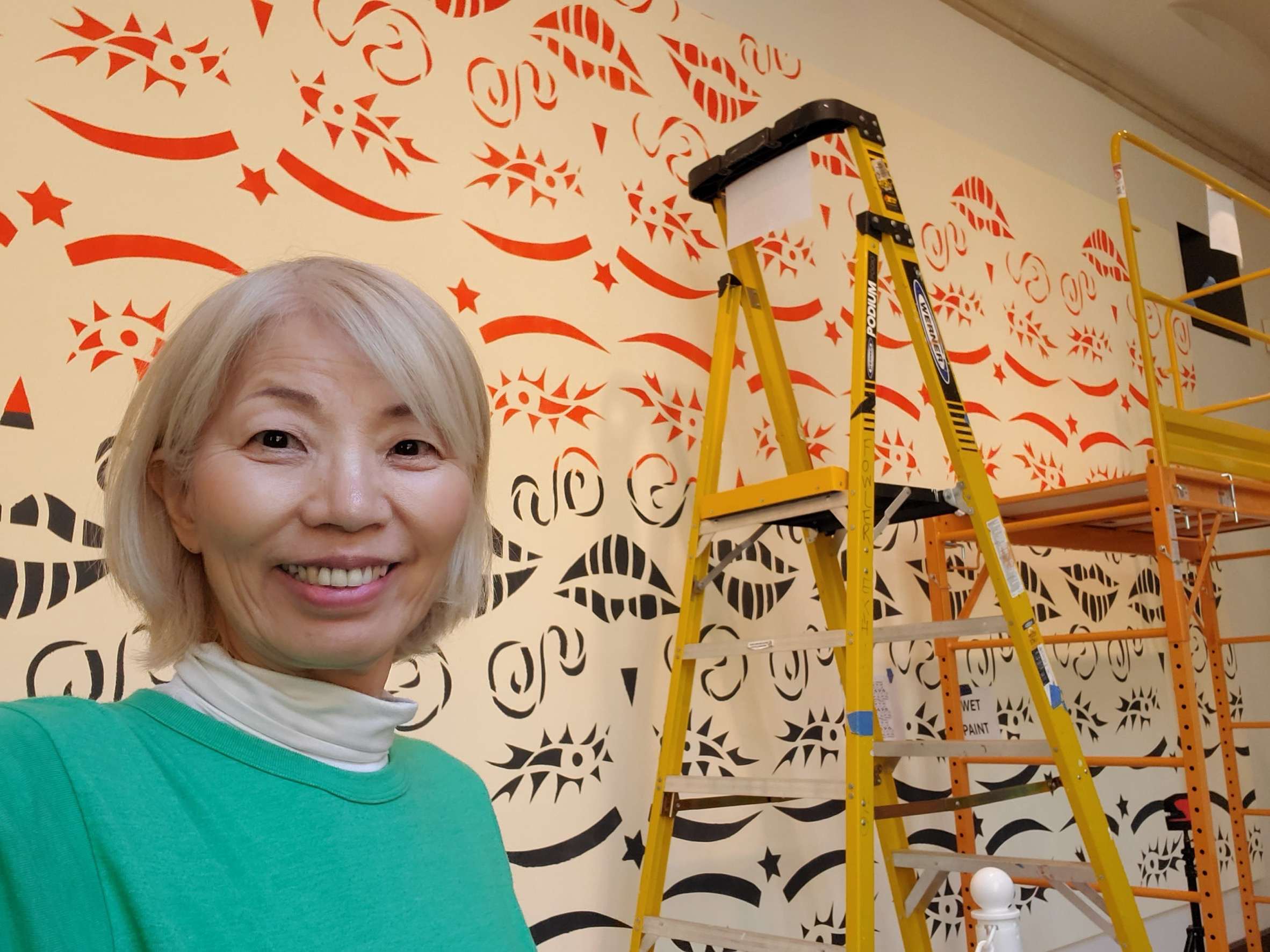 A woman takes a selfie in front of a mural with red and black symbols and shapes