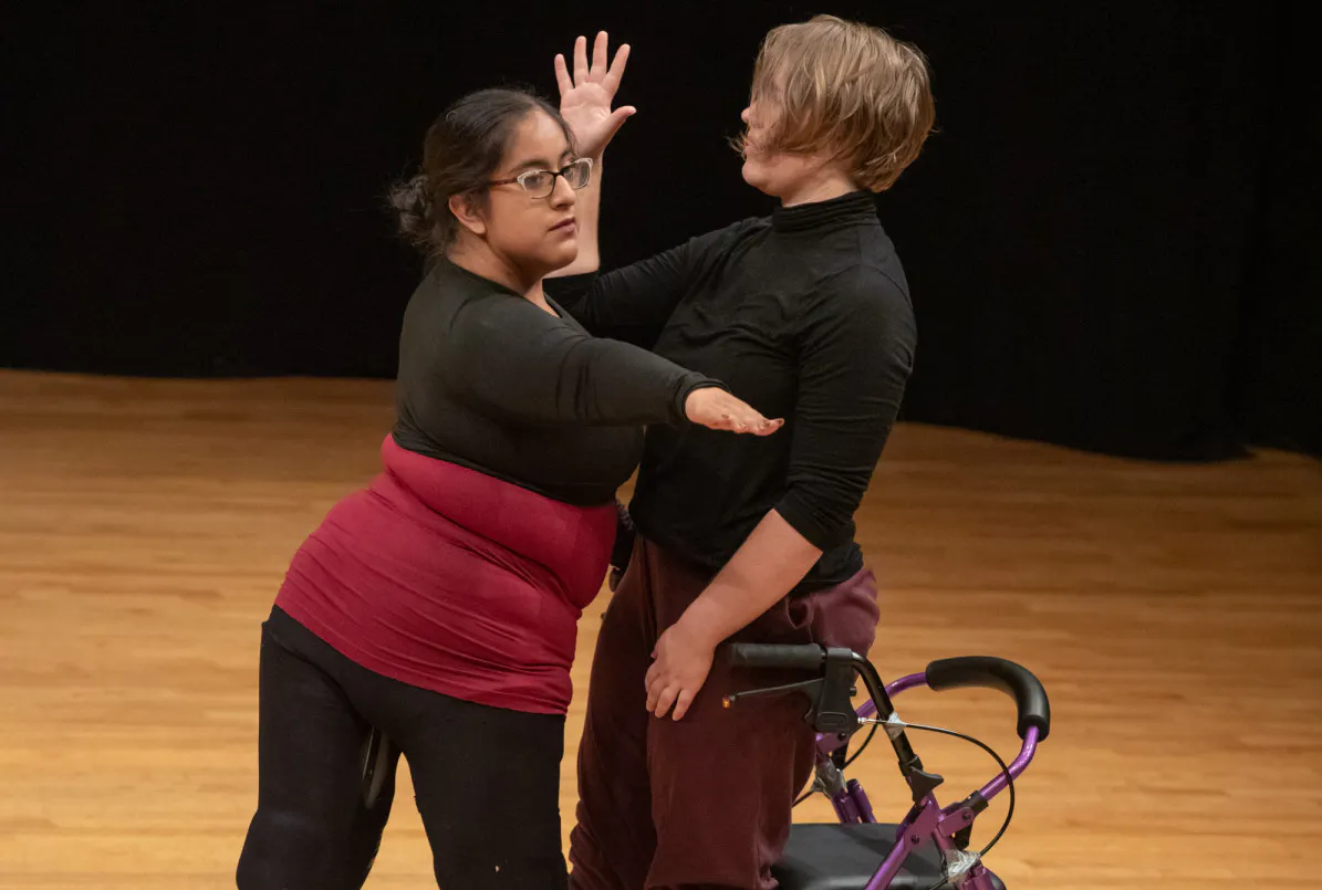 Two dancers face one another, standing closely together. A light skinned dancer (Bailey Anderson), dressed in a black turtleneck and sweats, raises her right arm, palm facing forward. A brown skinned dancer, Vanessa Nightsky Cruz, dressed in black leggings and a burgundy and black top looks out over her extended right arm, which stretches to her side, palm down. A mobility device, (AKA Pluto) rests behind Bailey.