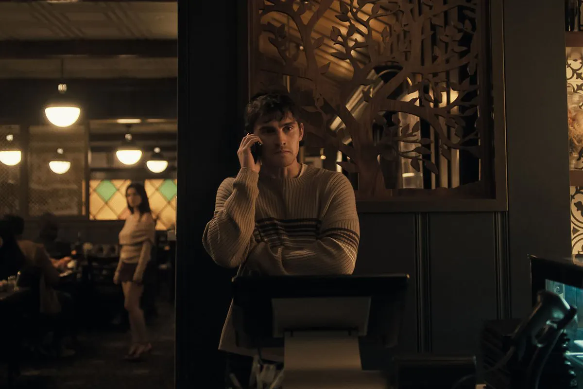 A young man in a striped sweater holds a cell phone next to his head, while out-of-focus in the background, a long-haired figure in a beige sweater and dark skirt looks his way