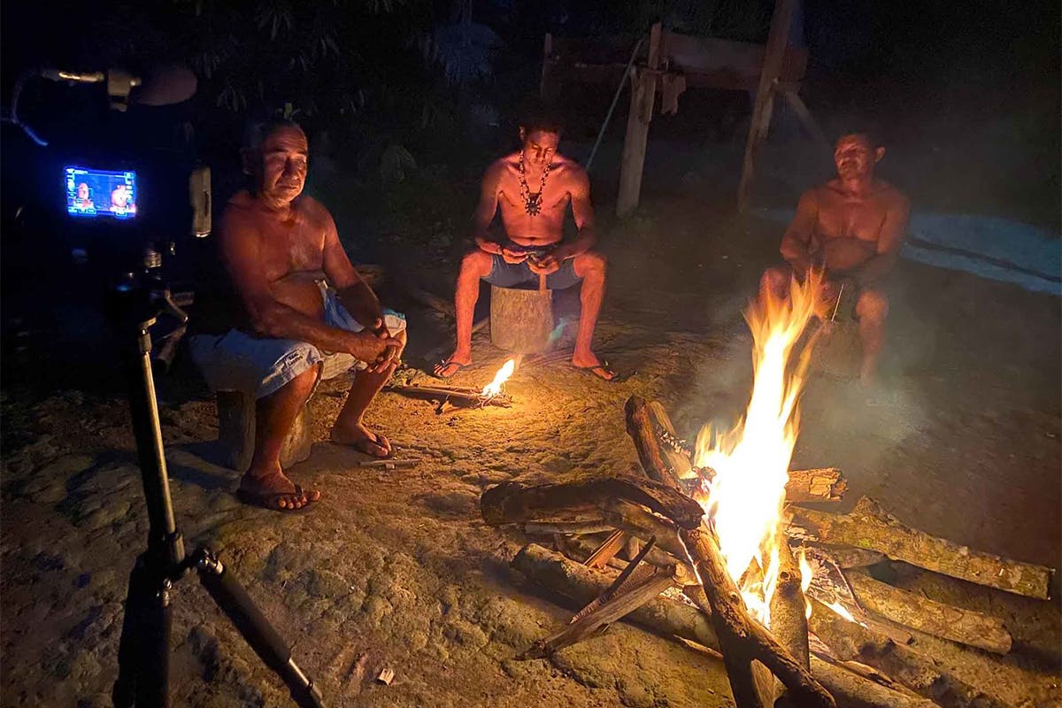 Three men sit in the dark around a fire. A camera captures their discussion. A screen shot from the film Stepping Softly on the Earth. 