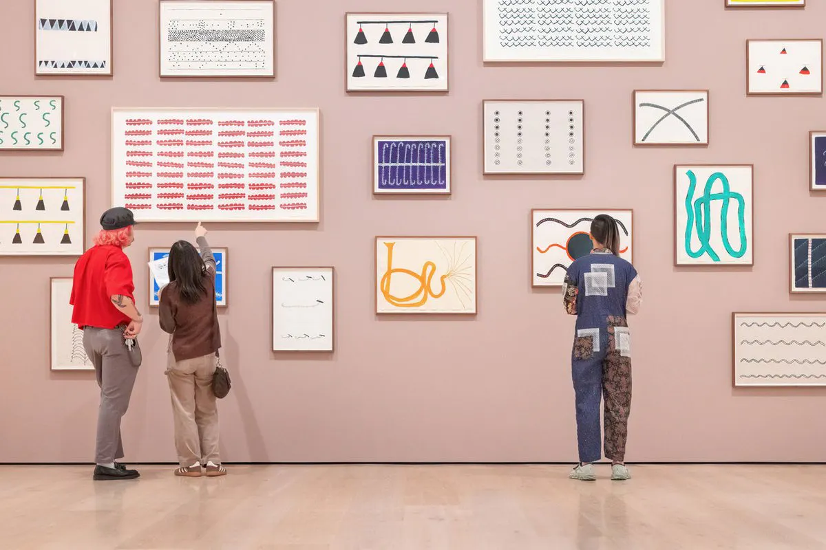 Three visitors, viewed from behind, stand in front of a pink gallery wall covered in a cluster of framed abstract artworks