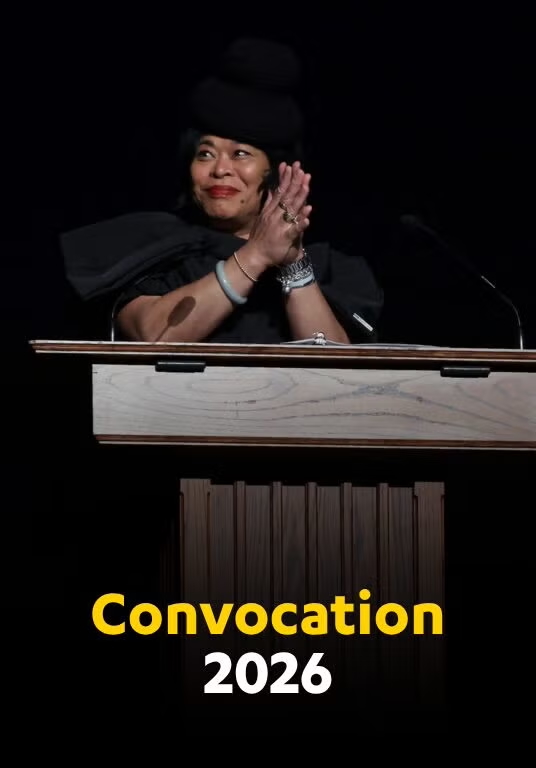 A woman in a black hat and dress stands at a lectern on stage, clapping her hands together. Text on the bottom of the image reads "Convocation 2026"