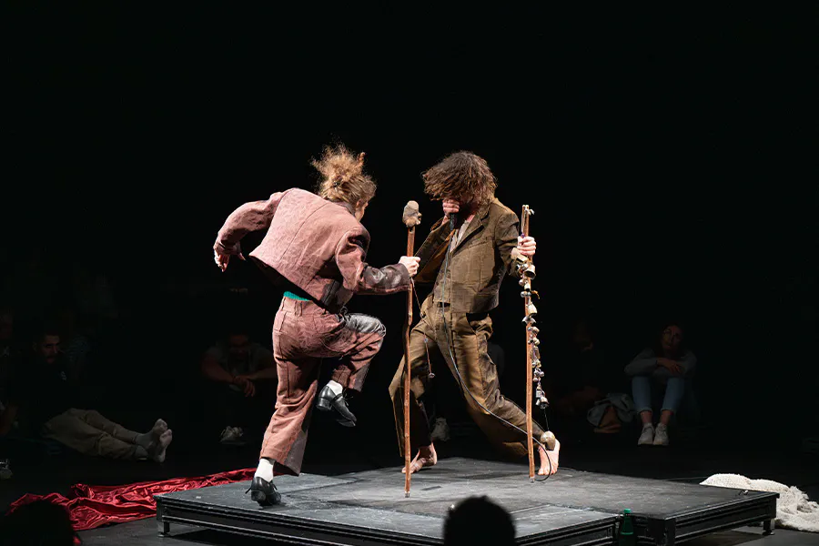 Performance image from Mirlitons featuring Hainaux and Chaignaud performing in the center of a theater on a small square stage. Silhouettes of audience members are seen in the foreground and background. Chaignaud faces away from the camera as he lifts on leg in the air, tap dancing. He wears pink pants and a jacket, holding a large wooden stick. Hainaux faces towards the camera and Chaignaud, wearing a brown suit and holding a wooden stick with bells attached to it. He beatboxes into a microphone.