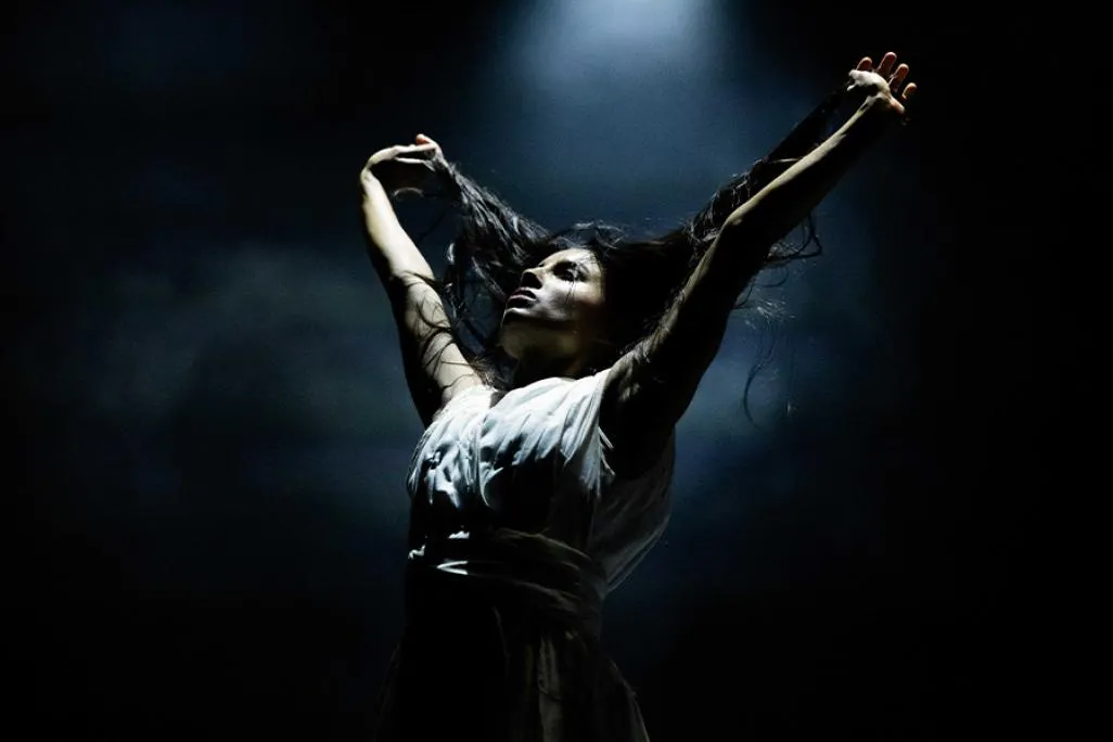 A dancer poses with arms raised above their head on a stage with a dark backdrop and smoke floating in the air.