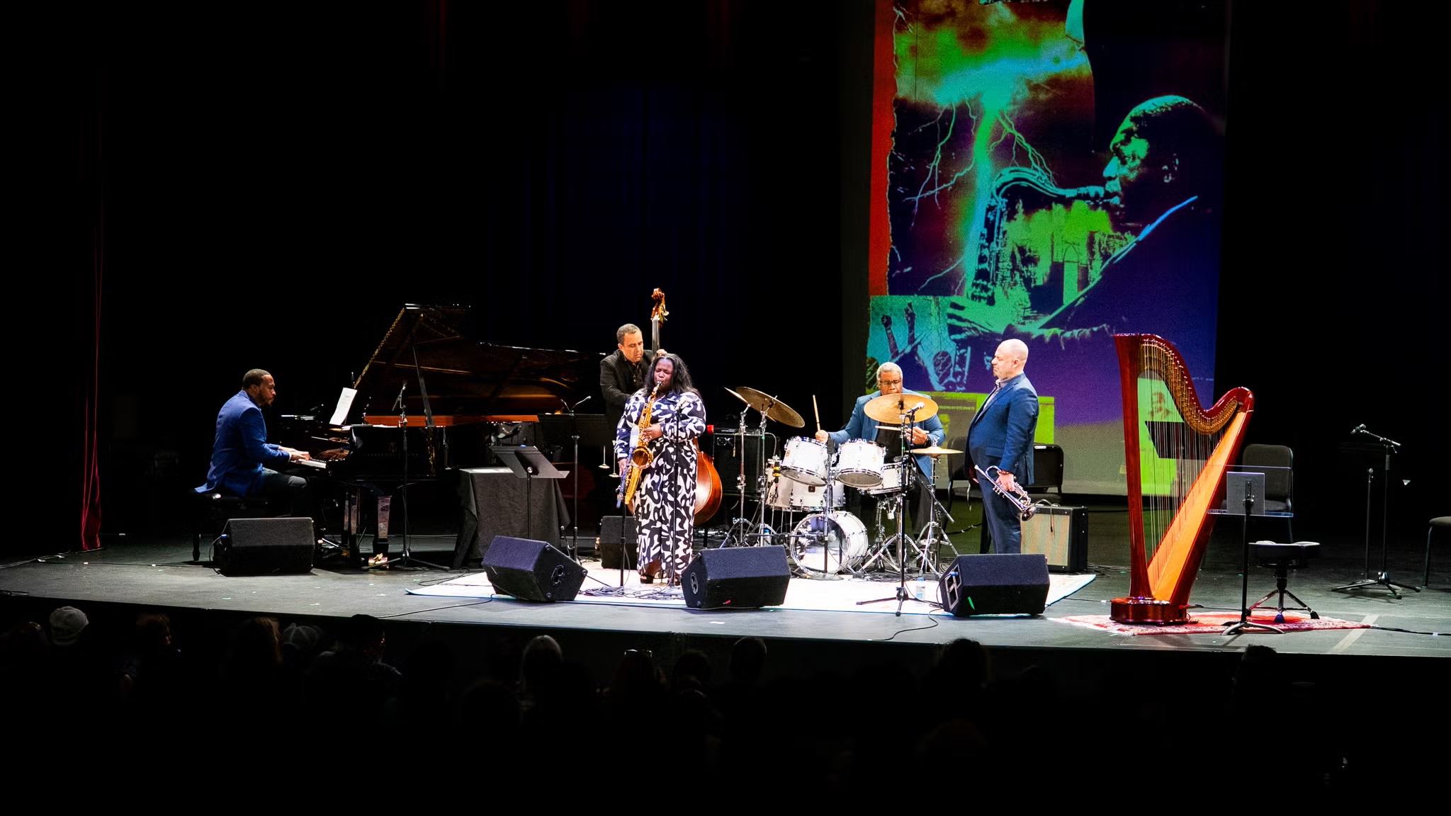 A group of jazz performers on stage with a large screen showing John Coltrane playing sax