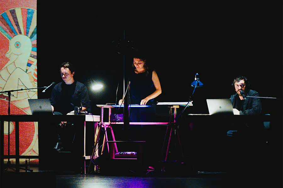 Cuervo performers seated and standing at illuminated workstations with laptops and microphones during a multimedia live performance on a darkened stage.