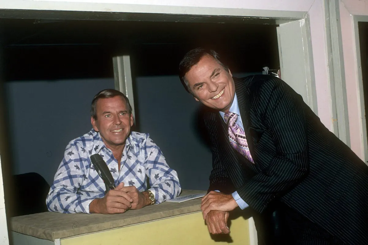 2 men smiling. one is sitting and one is standing leaning on a table