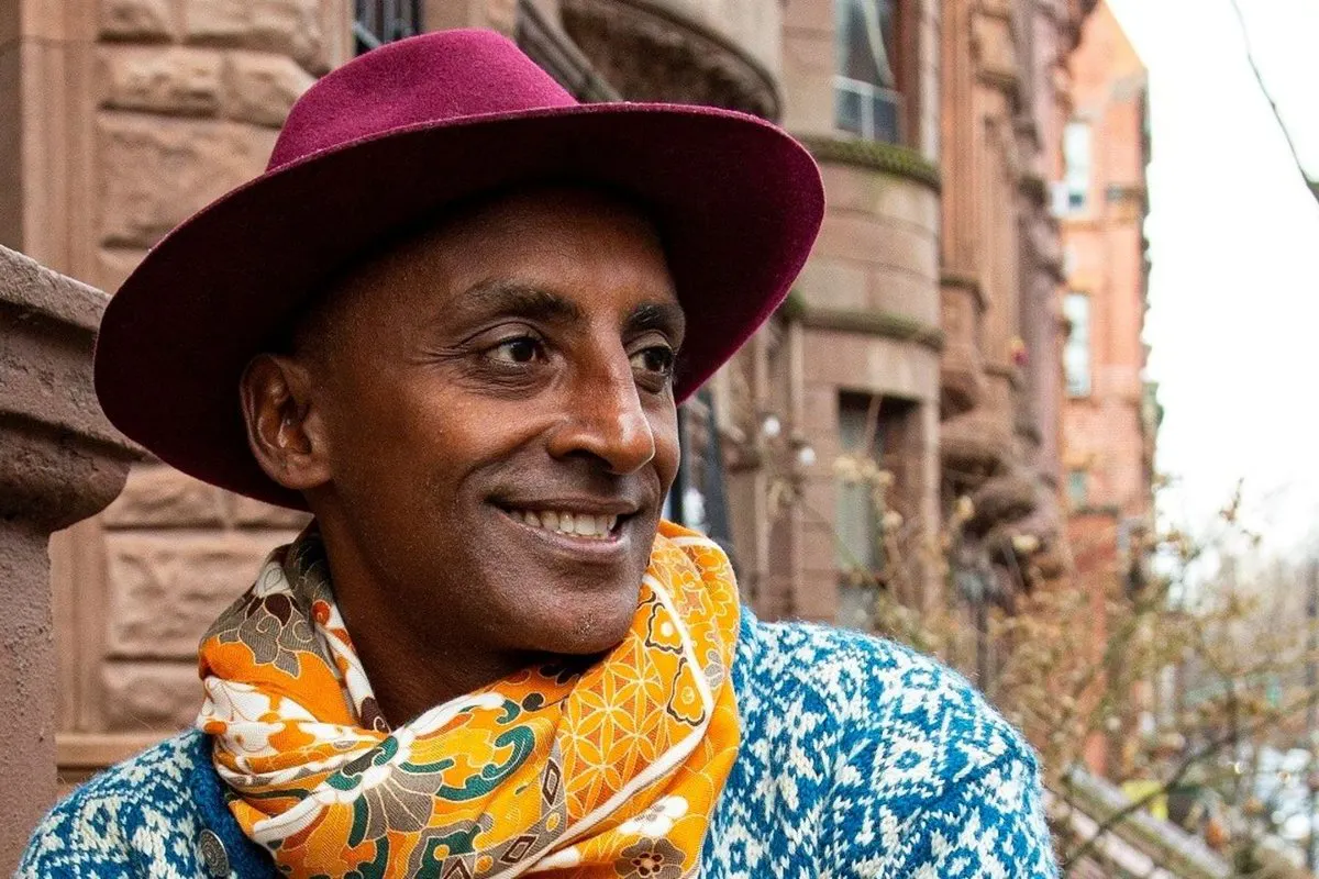 Smiling person in a burgundy hat, colorful patterned scarf, and blue-and-white sweater standing beside a brick building.