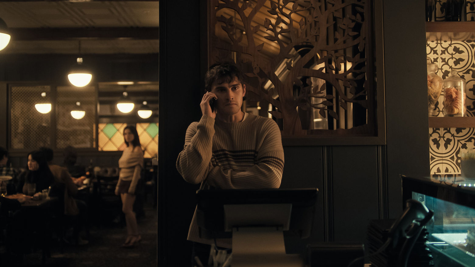 A scene from the film Obsession. A young man stands in the foreground of a restaurant, he is talking on the phone. 