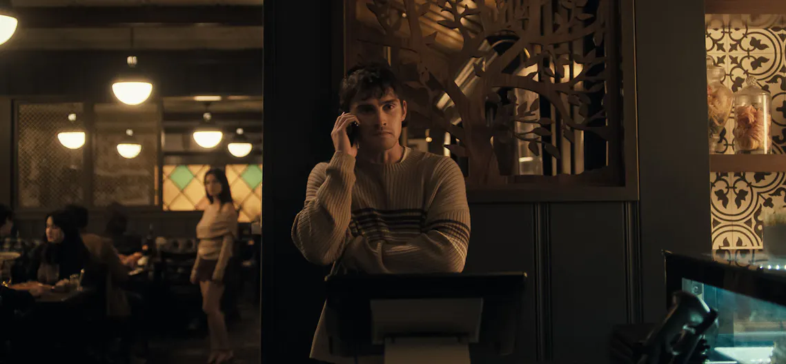 A scene from the film Obsession. A young man stands in the foreground of a restaurant, he is talking on the phone.