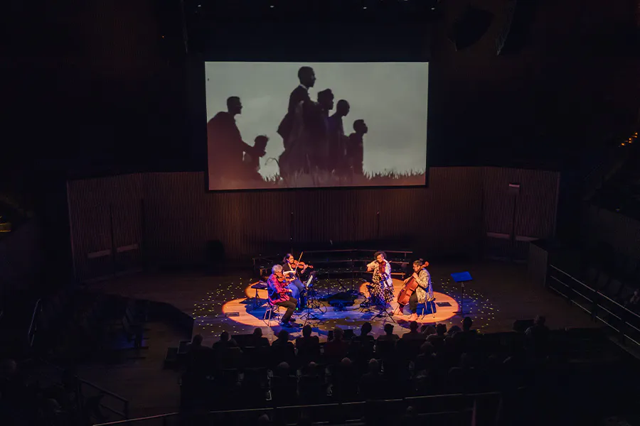 birds eye view of Kronos quartet members performing on stage amongst an audience with a projection in the background