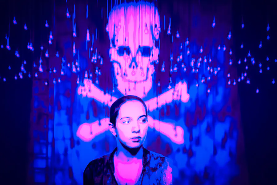 A woman stands in front a projection of a skull and bones, cast onto a sheet, illuminated by purple and blue light.