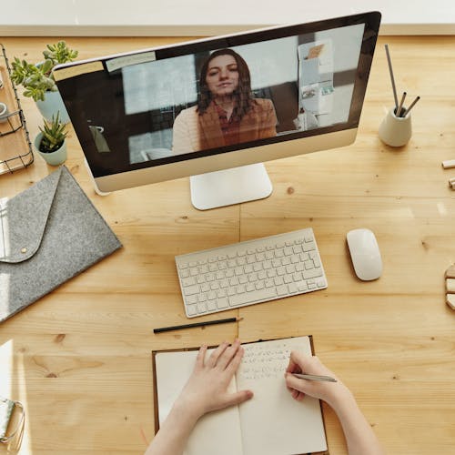 Women talking on a computer screen video conference call, while another woman is writing in a notebook on a wooden desk.