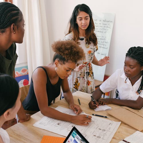Group of people sitting at a desk, with woman standing in front of a whiteboard.