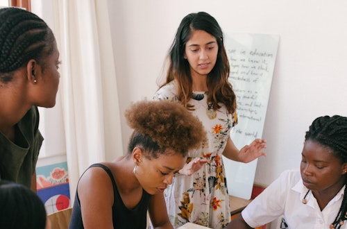 Group of people sitting at a desk, with woman standing in front of a whiteboard.