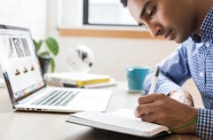Man in blue and white checked shirt writing in a notebook in front of a laptop.
