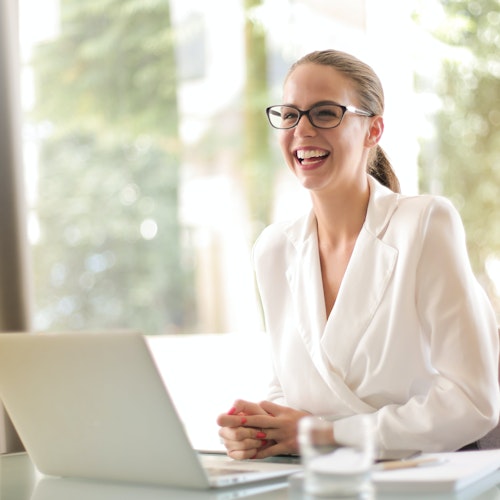 Woman wearing a white suit smiling and sitting at her laptop looking engaged