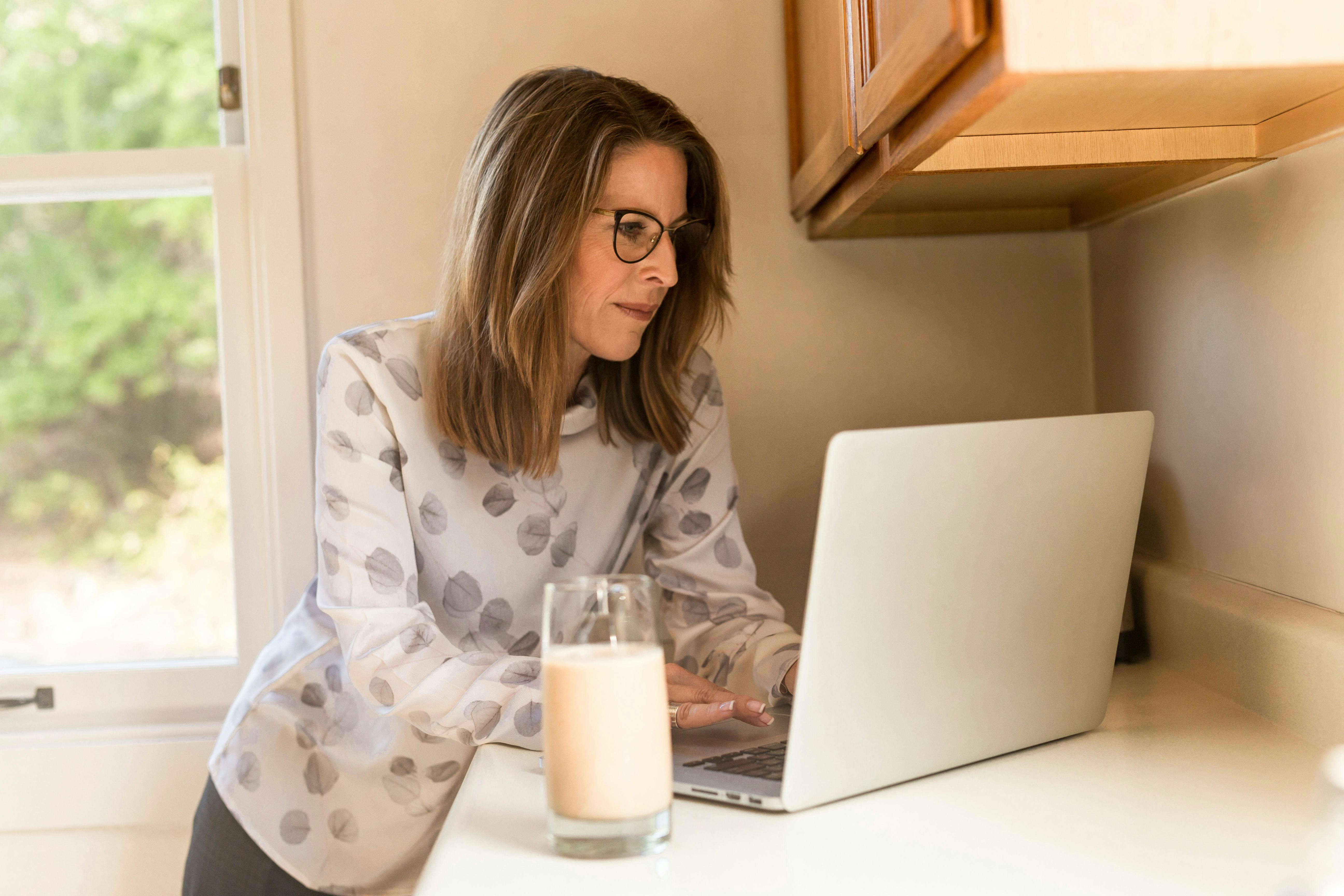 Woman standing at her kitchen bench looking at a laptop and drinking a glass of milk