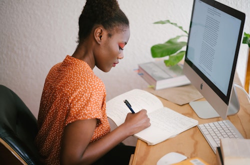 Woman at a desk writing in a notepad in front of a computer. 