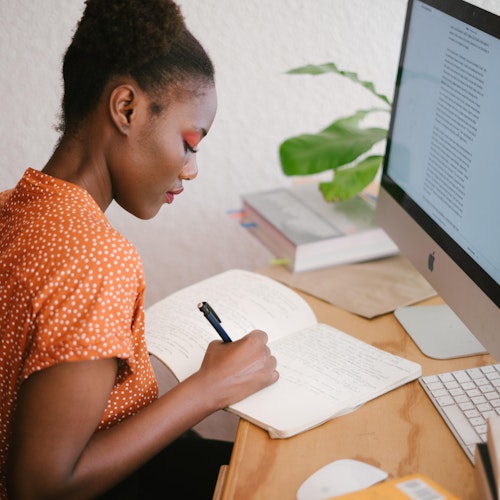 Woman at a desk writing in a notepad in front of a computer. 