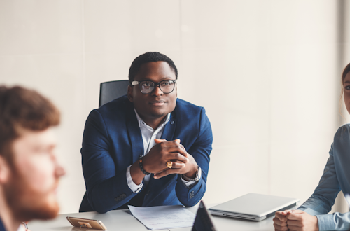 Man sitting at the head of a long office table, listening to a presentation