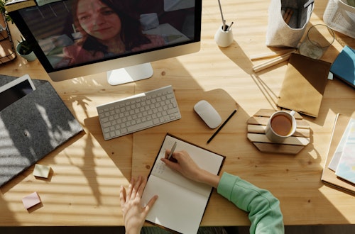 Woman sitting at her desk writing in her notepad, talking to another woman on screen via Zoom.